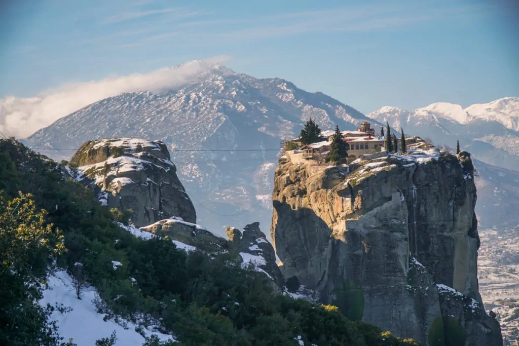 Snow-covered Meteora monastery on top of rocky cliffs with mountains in the background, Greece winter landscape