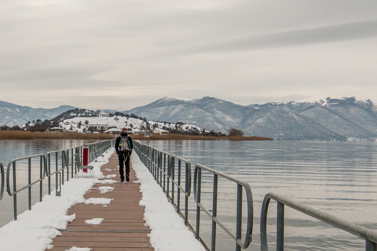 Traveler walking on wooden pier by Prespes Lake with snowy mountains in the background, Northern Greece winter scenery